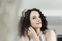 hair styling for wedding - bride putting on earrings with curly dark brown hair
